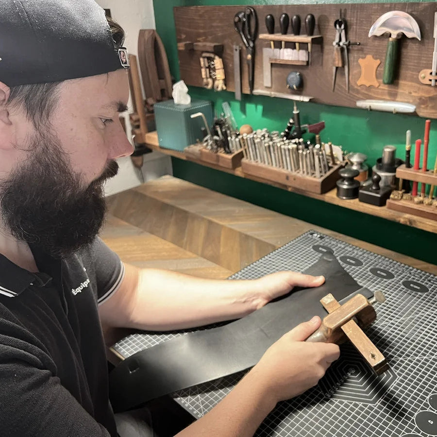 Artisan using a traditional wooden strap cutter to slice black vegetable-tanned leather hide over a grid cutting mat in the Deco Leather Studio workshop.