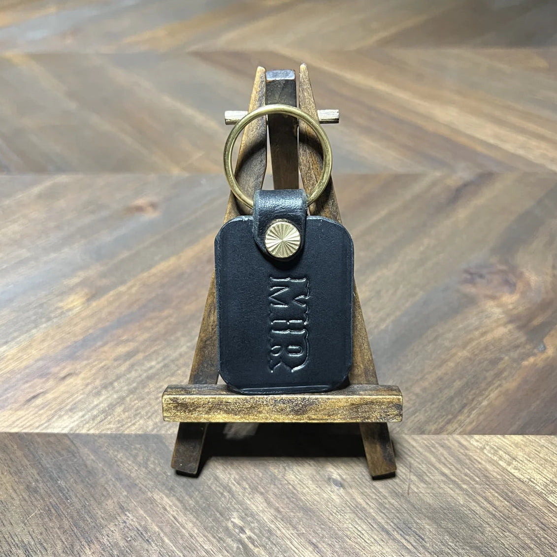 Handcrafted black vegetable-tanned leather key fob with custom "MR" stamped monogram and solid brass split ring resting on a mini wooden display easel over a chevron oak desk at Deco Leather Studio in Miami Beach.