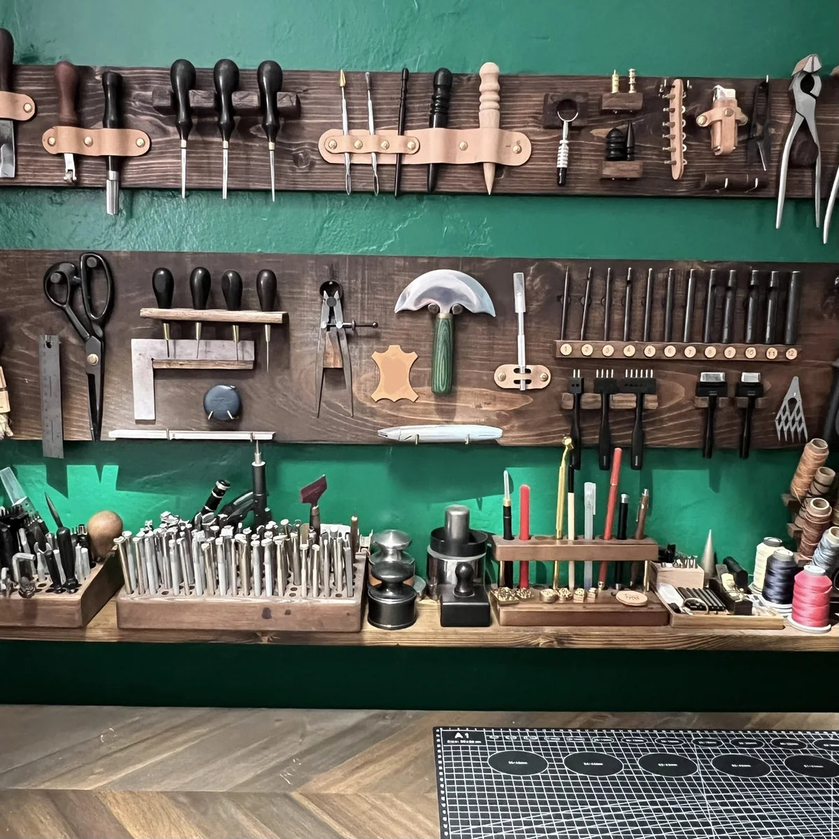 Close-up view of traditional leatherworking hand tools including French skivers, stitching chisels, edge bevelers, and brass stamps lined up along a wooden workbench shelf at Deco Leather Studio.