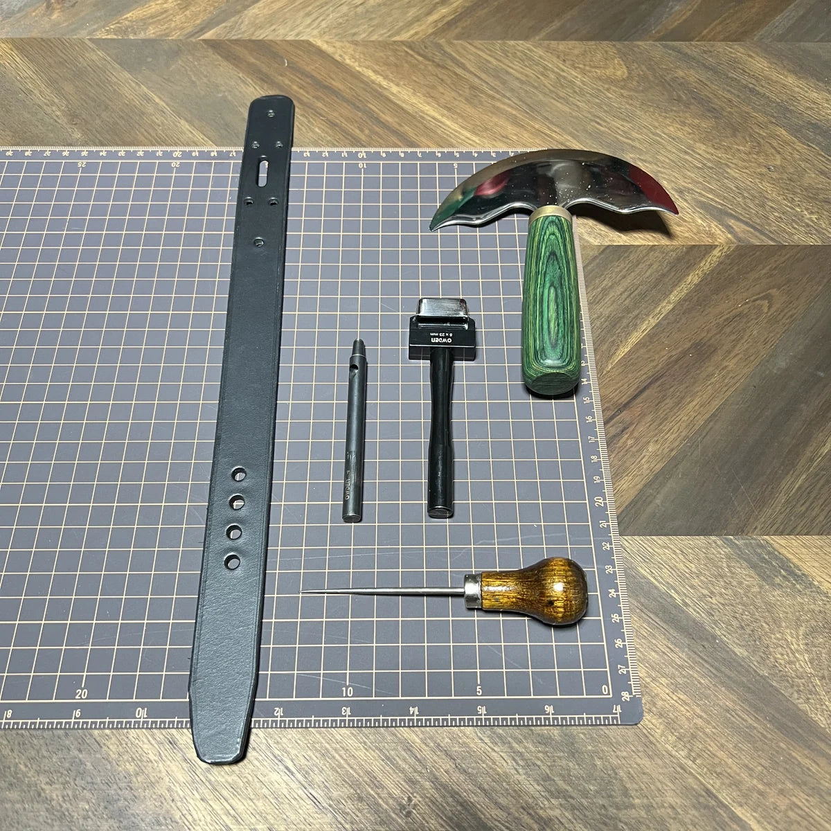Precision leathercraft benchwork setup featuring a cut black leather cuff strap, green-handled head knife, round punch, oblong slot punch, and scratching awl arranged on a gridded cutting mat at Deco Leather Studio.