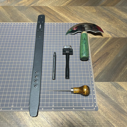 Precision leathercraft benchwork setup featuring a cut black leather cuff strap, green-handled head knife, round punch, oblong slot punch, and scratching awl arranged on a gridded cutting mat at Deco Leather Studio.