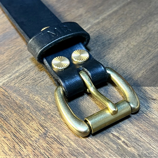 Close-up detail of a handcrafted black vegetable-tanned leather cuff bracelet featuring solid brass buckle hardware, decorative starburst rivets, and a custom "MR" stamped keeper loop on a chevron oak workbench at Deco Leather Studio in Miami Beach.