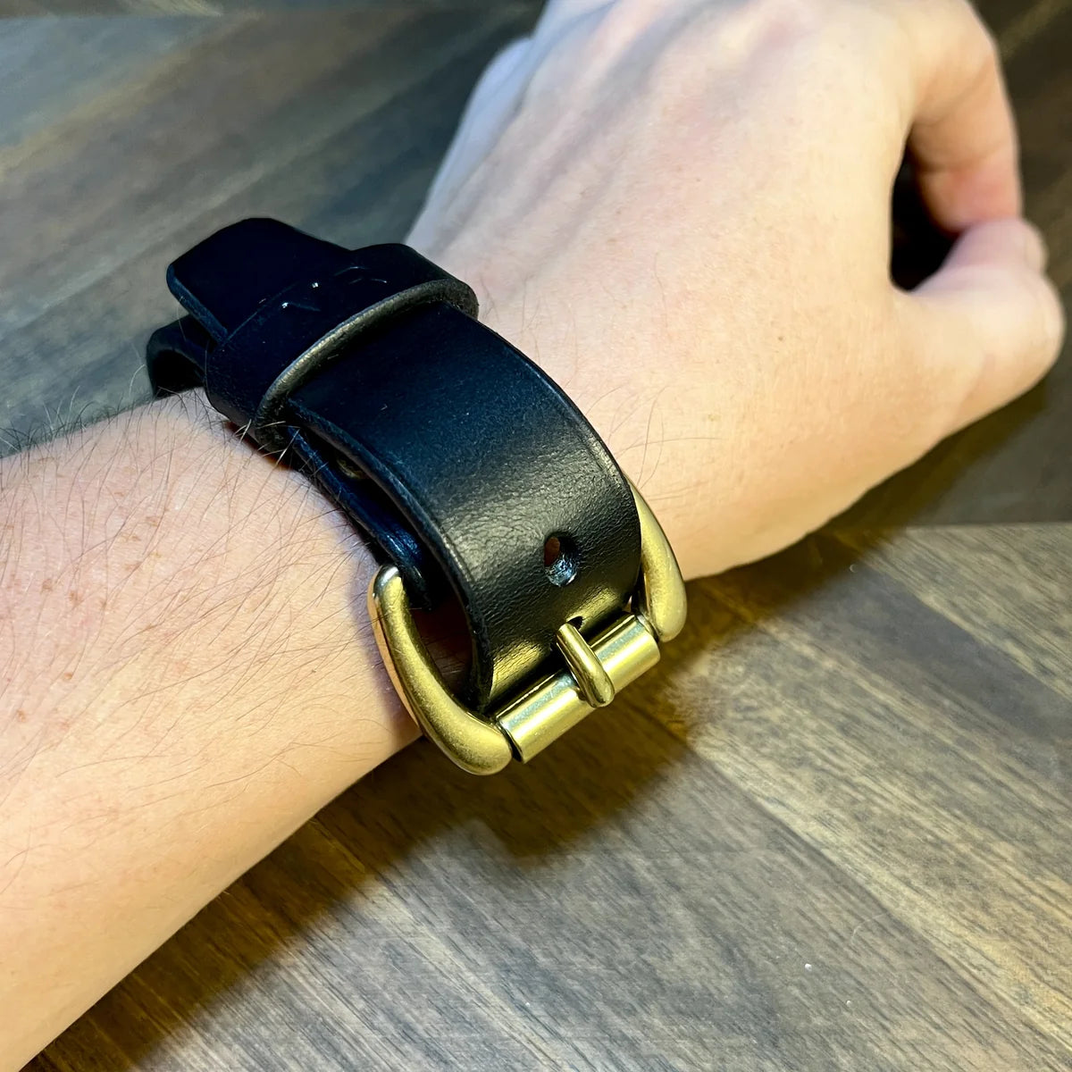 Handcrafted black vegetable-tanned leather cuff bracelet with solid brass roller buckle and hot-stamped keeper loop worn on a wrist over a chevron oak workbench at Deco Leather Studio in Miami Beach.