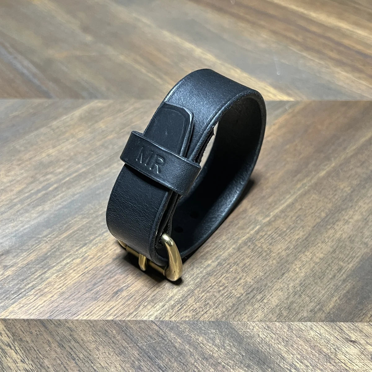 Handcrafted black vegetable-tanned leather cuff bracelet with custom "MR" stamped monogram keeper loop and solid brass buckle, buckled into a curved roll on a chevron oak workbench at Deco Leather Studio in Miami Beach.