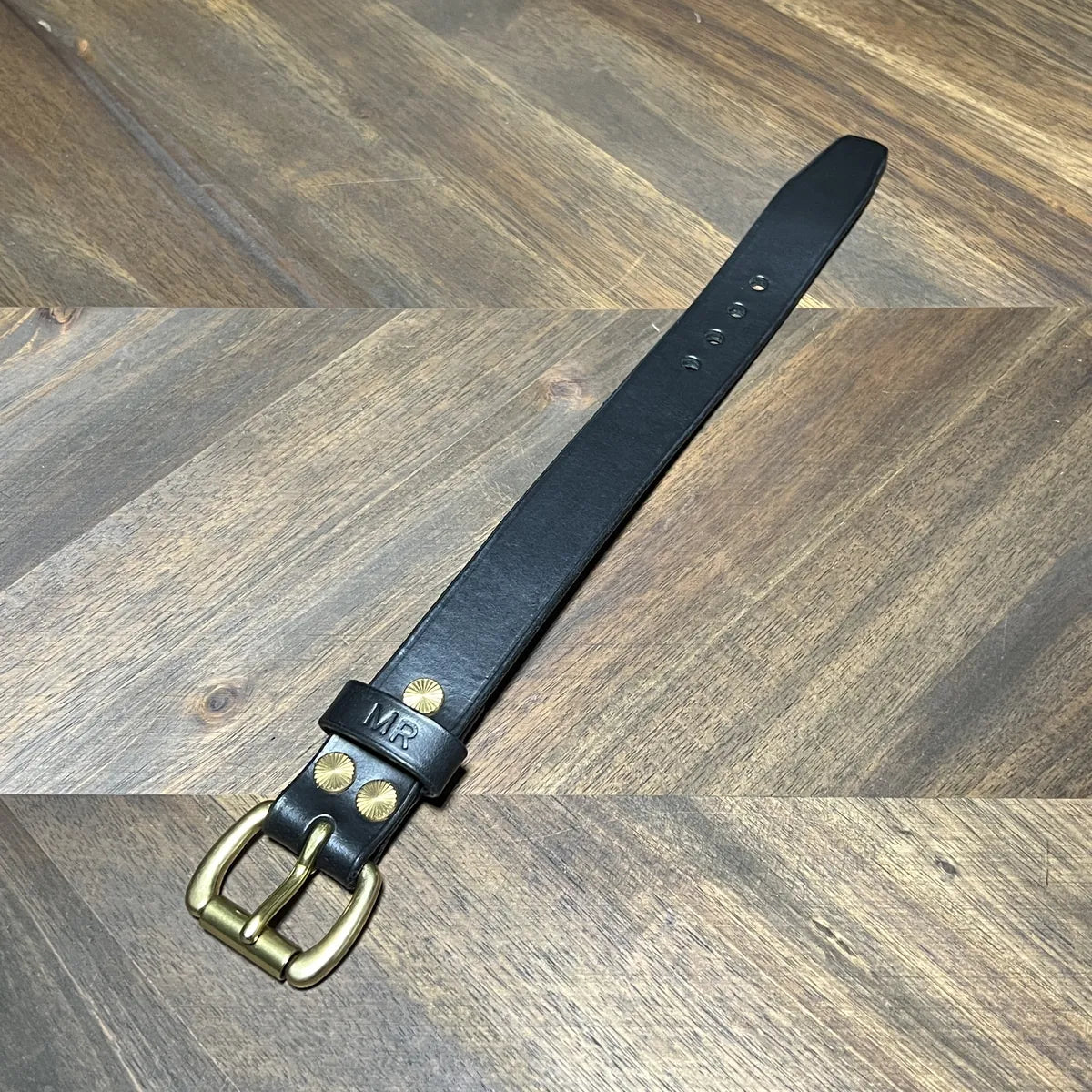 Handcrafted black vegetable-tanned leather cuff bracelet laid flat showing solid brass buckle, decorative starburst brass rivets, custom "MR" monogram keeper, and sizing holes over a chevron oak desk at Deco Leather Studio in Miami Beach.