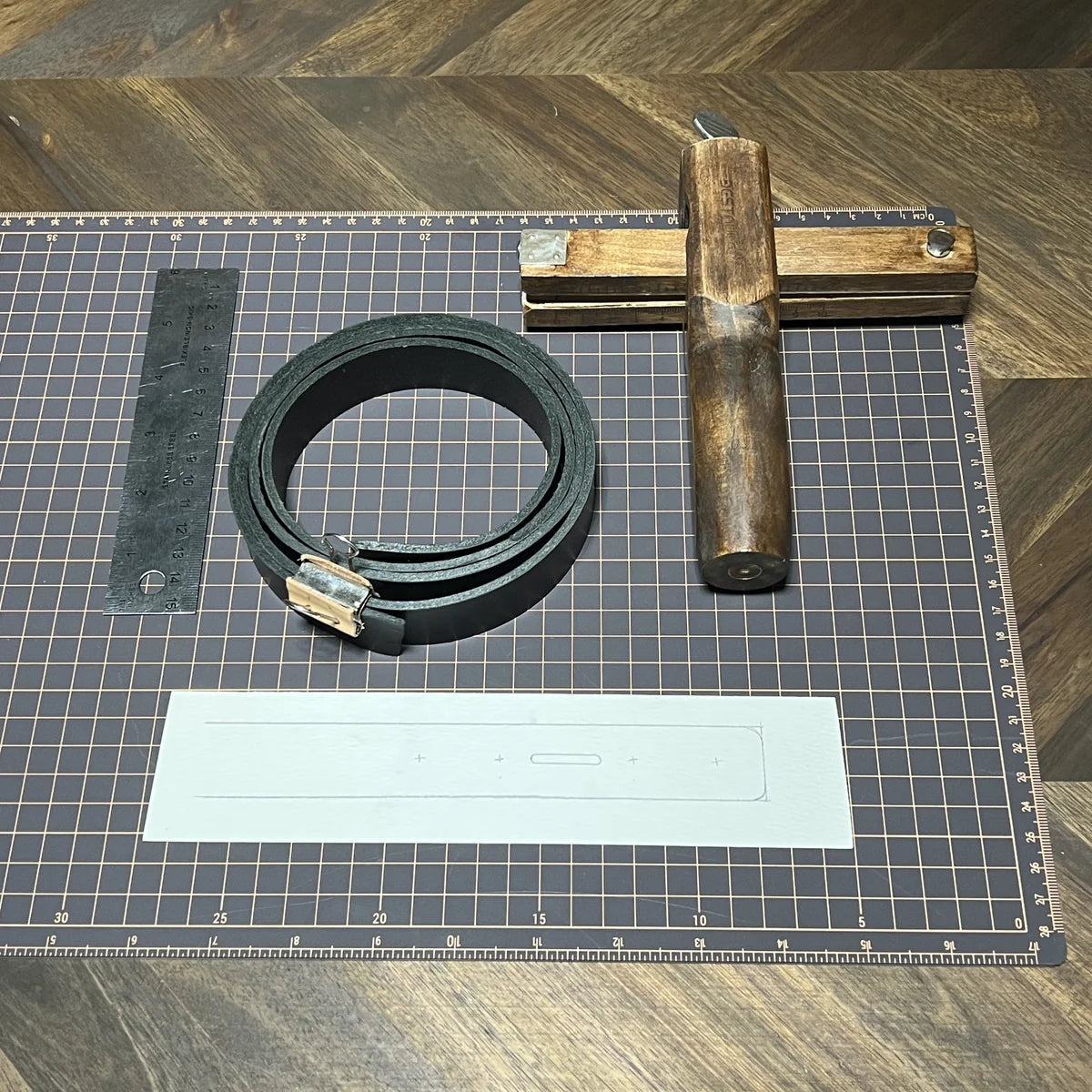 Raw material preparation showing a coiled 8oz black vegetable-tanned leather strap, wooden plough gauge strap cutter, stainless steel ruler, paper template, and gridded cutting mat at Deco Leather Studio.