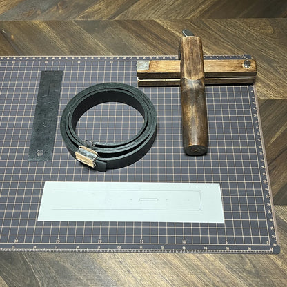 Raw material preparation showing a coiled 8oz black vegetable-tanned leather strap, wooden plough gauge strap cutter, stainless steel ruler, paper template, and gridded cutting mat at Deco Leather Studio.