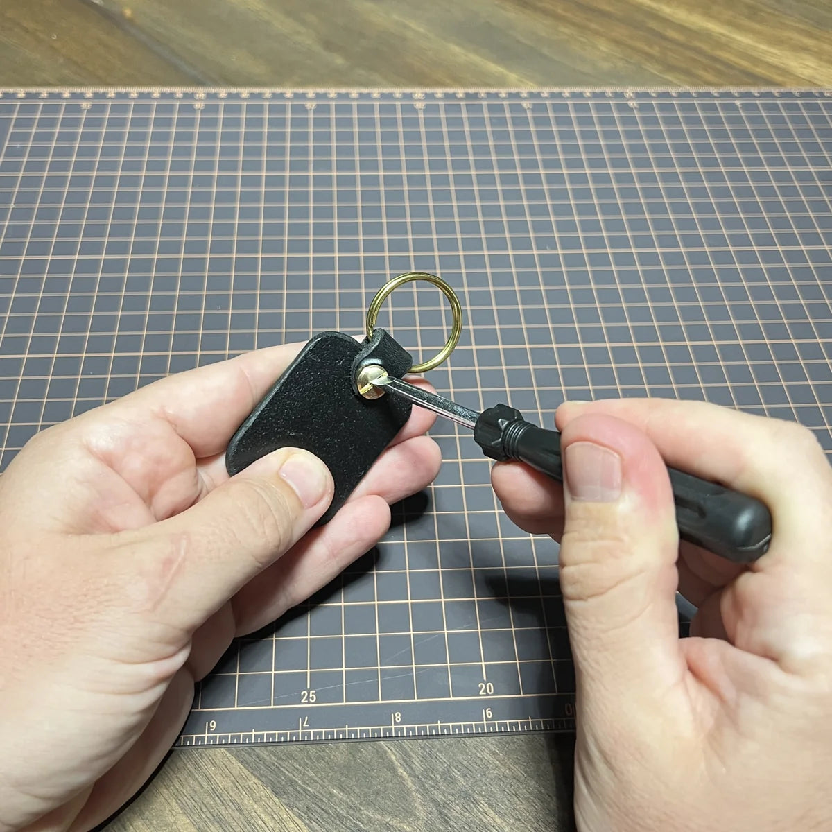 Craftsperson using a small flathead screwdriver to tighten a solid brass Chicago screw on a black vegetable-tanned leather key fob with a brass split ring over a gridded cutting mat at Deco Leather Studio in Miami Beach.