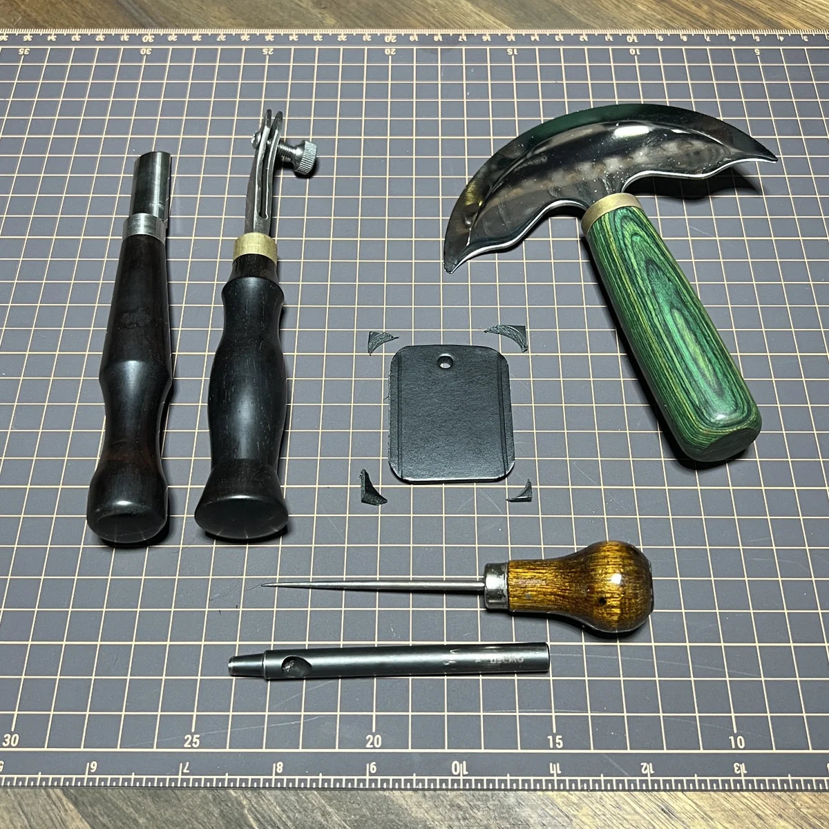 Precision leathercraft benchwork setup featuring green-handled head knife, edge bevelers, scratching awl, and drive punch around a cut 8oz leather key fob blank on a gridded cutting mat.