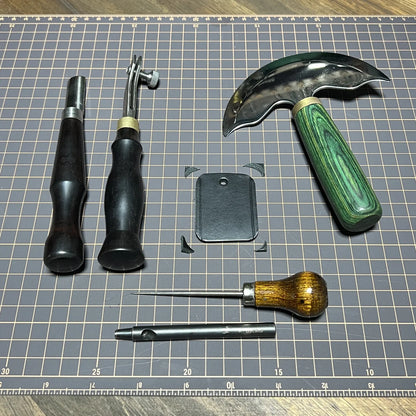 Precision leathercraft benchwork setup featuring green-handled head knife, edge bevelers, scratching awl, and drive punch around a cut 8oz leather key fob blank on a gridded cutting mat.