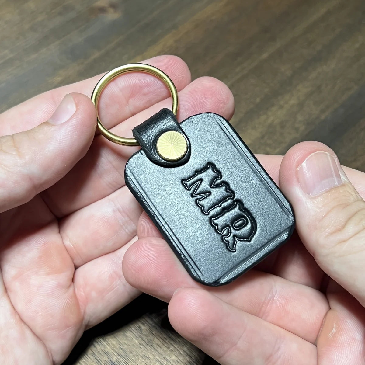 Close-up held in hands displaying a handcrafted black vegetable-tanned leather key fob featuring custom "MR" hot-stamped monogram, brass split ring, and sunburst rivet hardware at Deco Leather Studio in Miami Beach.