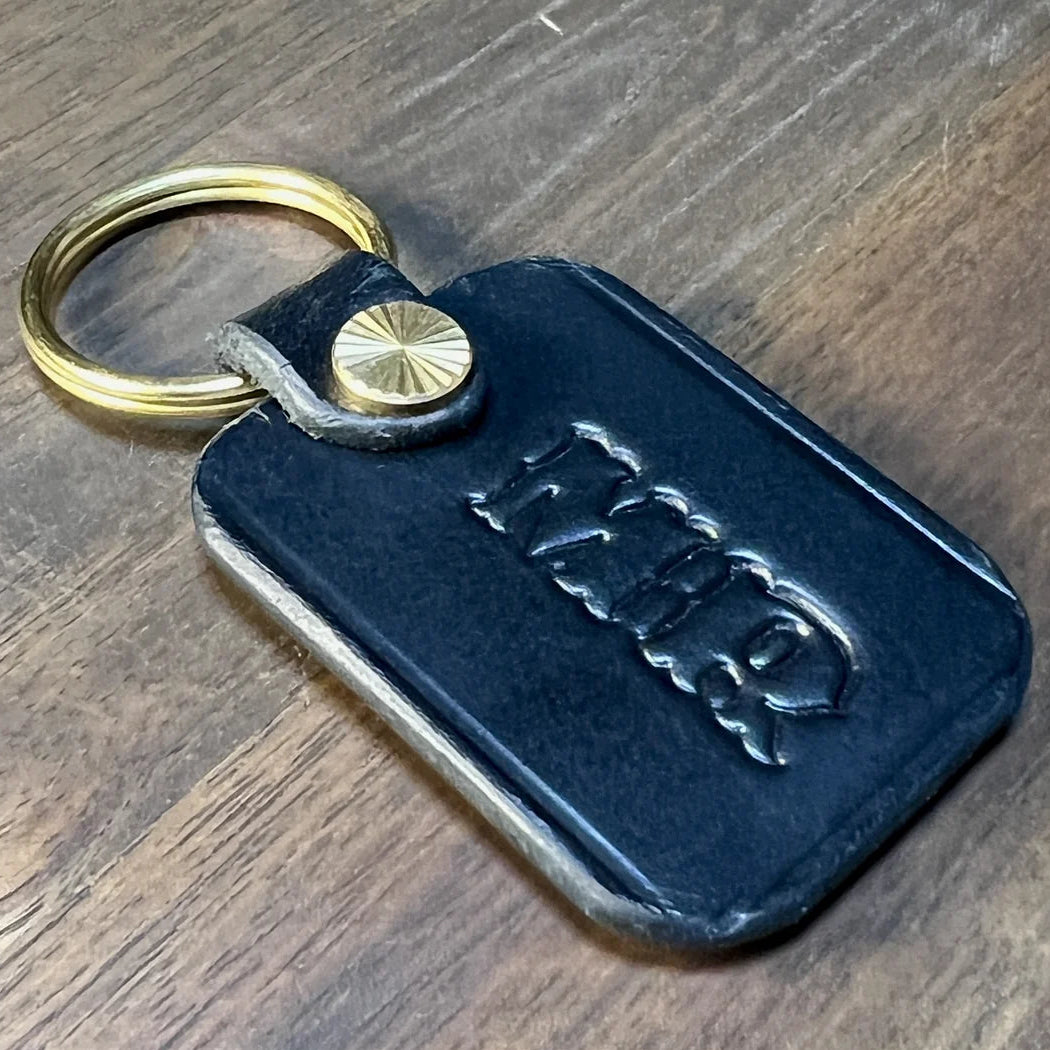 Handcrafted black vegetable-tanned leather key fob with custom "MR" stamped monogram, decorative edge grooving, and solid brass split ring with sunburst brass rivet resting on a dark stained oak surface at Deco Leather Studio in Miami Beach.