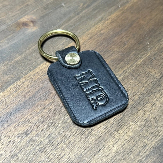 Handcrafted black vegetable-tanned leather key fob with custom "MR" hot-stamped monogram and solid brass split ring on dark oak workbench at Deco Leather Studio in Miami Beach.