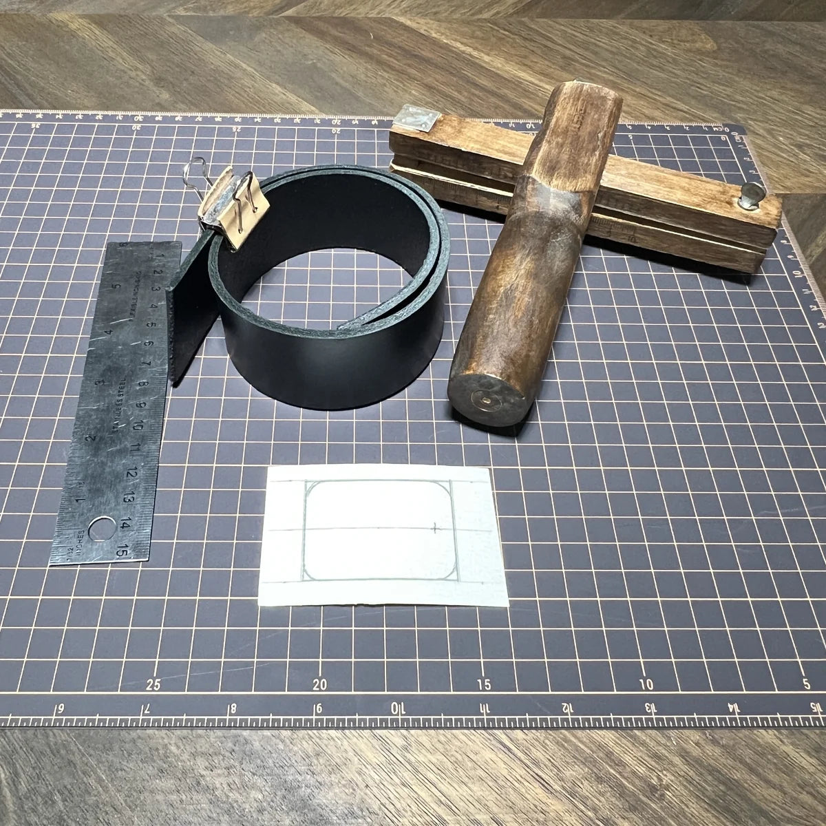 Raw material preparation including a roll of 8oz black veg-tan leather strip, wooden plough gauge strap cutter, steel ruler, paper template, and strap cutter.