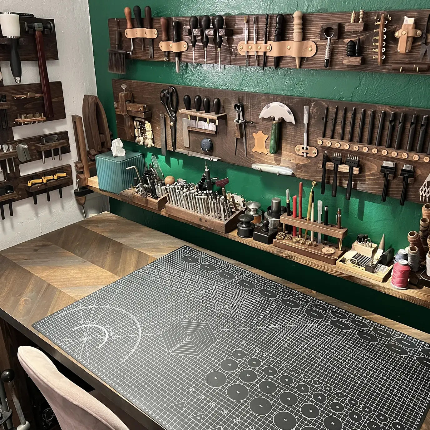 Wide-angle perspective of the main leathercraft workbench with grid cutting mat, herringbone oak desk surface, and custom wall tool organization at Deco Leather Studio.