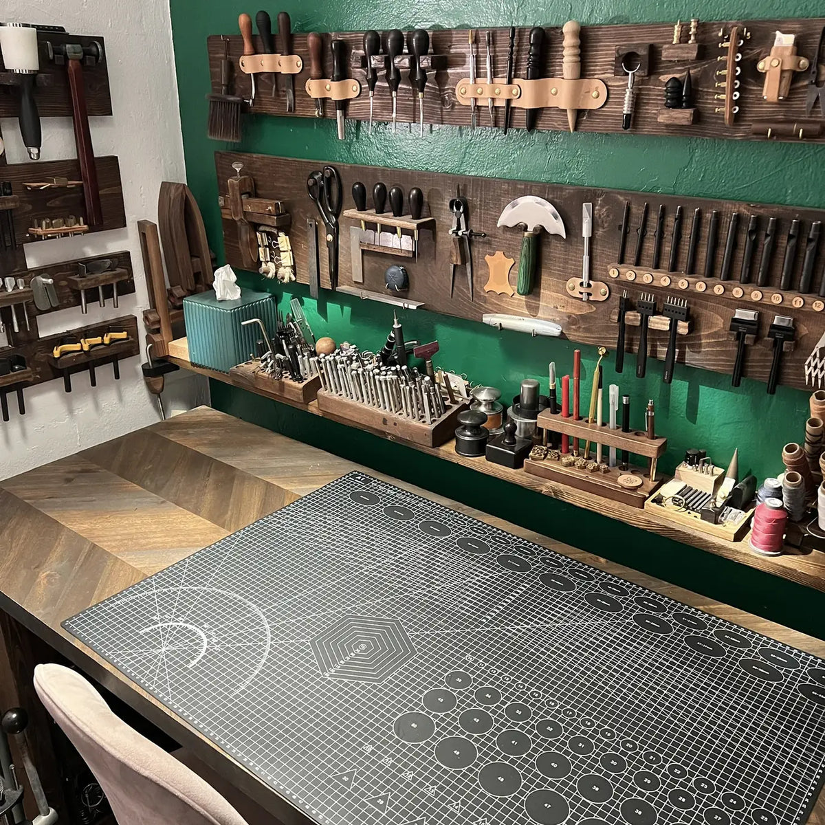 Wide-angle perspective of the main leathercraft workbench with grid cutting mat, herringbone oak desk surface, and custom wall tool organization at Deco Leather Studio.