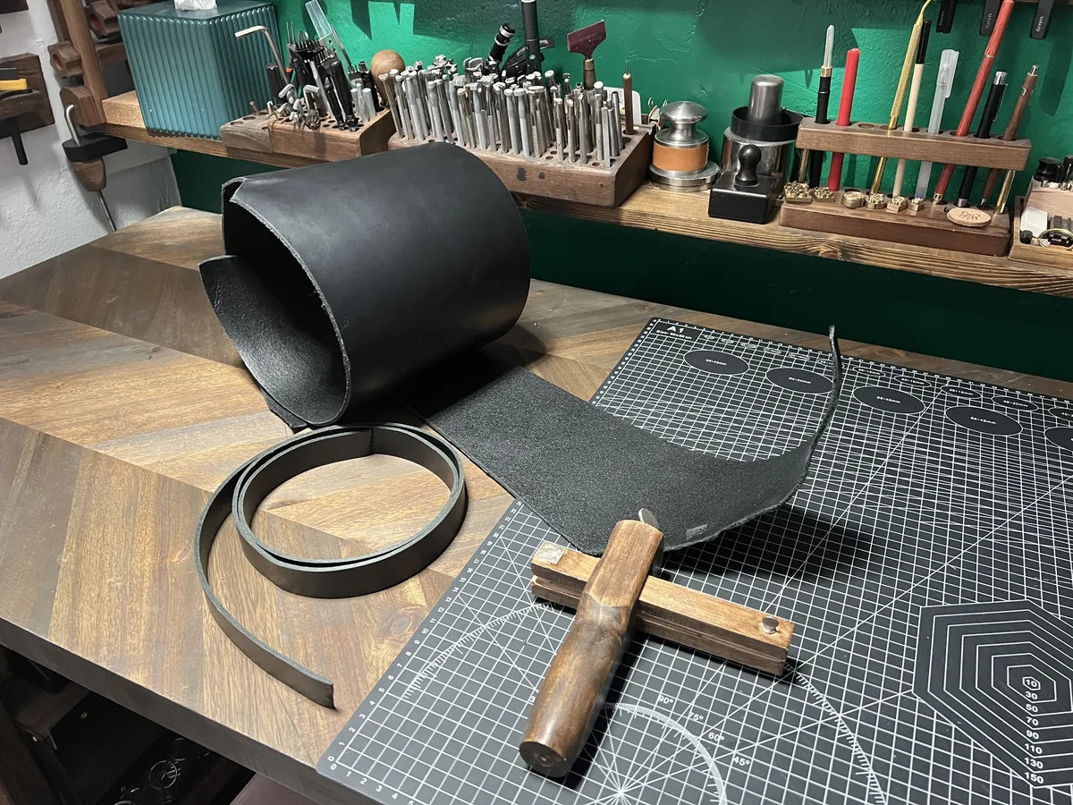 Rolled hide of 8 oz black vegetable-tanned leather alongside a wooden strap cutter and freshly cut 1.5" leather strip on a chevron wood workbench at Deco Leather Studio.
