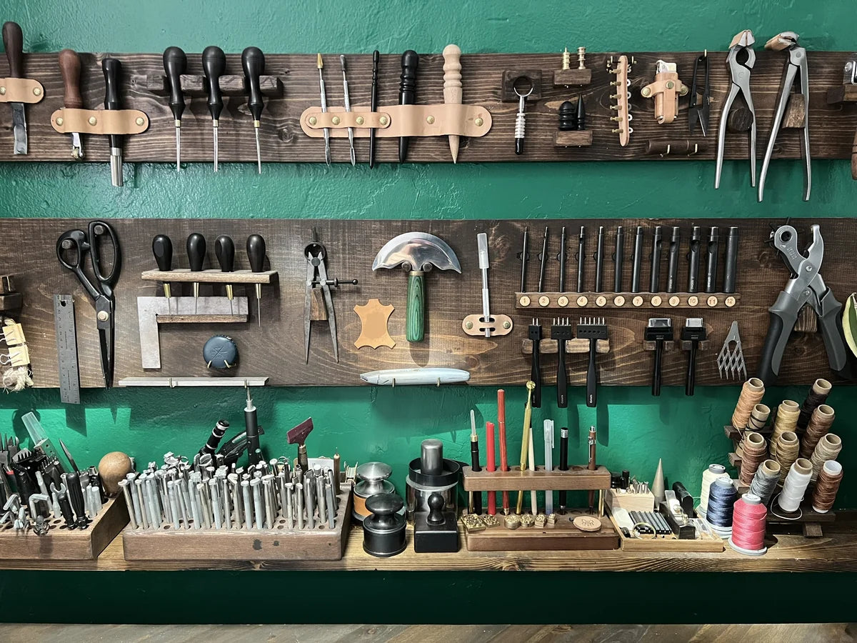 Organized wall-mounted dark wood tool rack featuring edge bevelers, pricking irons, round knives, and thread spools against an emerald green accent wall at Deco Leather Studio.