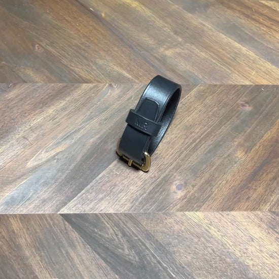 Silent 1080p product video loop demonstrating hands unbuckling a custom black vegetable-tanned leather cuff bracelet with hot-stamped "MR" monogram and solid brass buckle over a chevron oak workbench at Deco Leather Studio in Miami Beach.