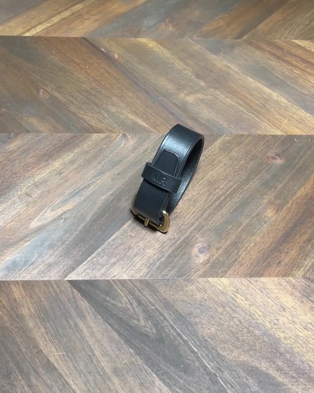 Silent 1080p product video loop demonstrating hands unbuckling a custom black vegetable-tanned leather cuff bracelet with hot-stamped "MR" monogram and solid brass buckle over a chevron oak workbench at Deco Leather Studio in Miami Beach.