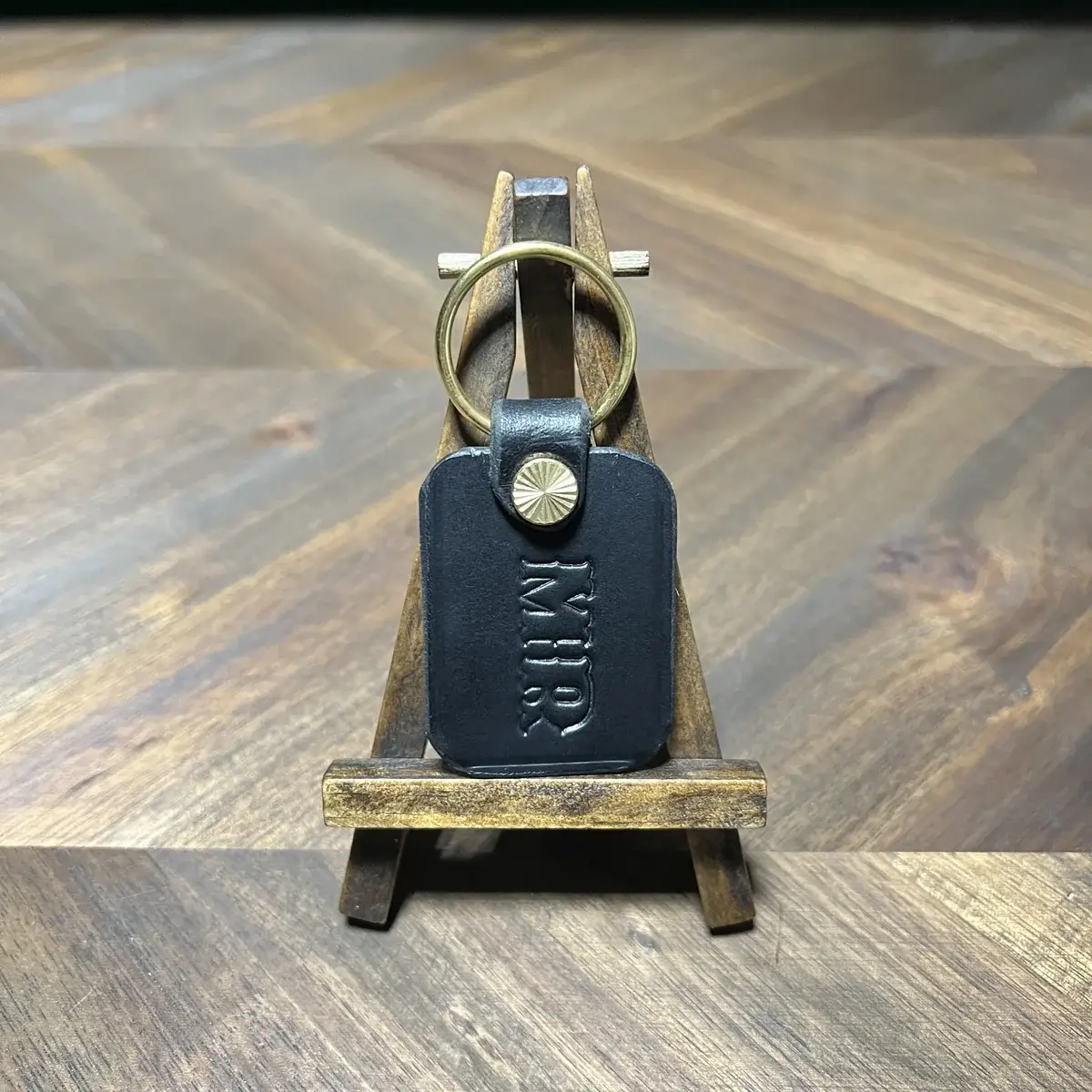 Black leather keychain with a brass ring and stamped initials on a small easel
